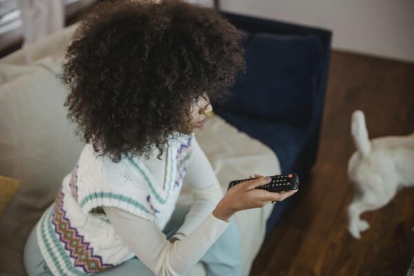 From above of focused young black woman with Afro hair in casual outfit sitting on sofa with remote controller in hand and watching TV during weekend at home with dog