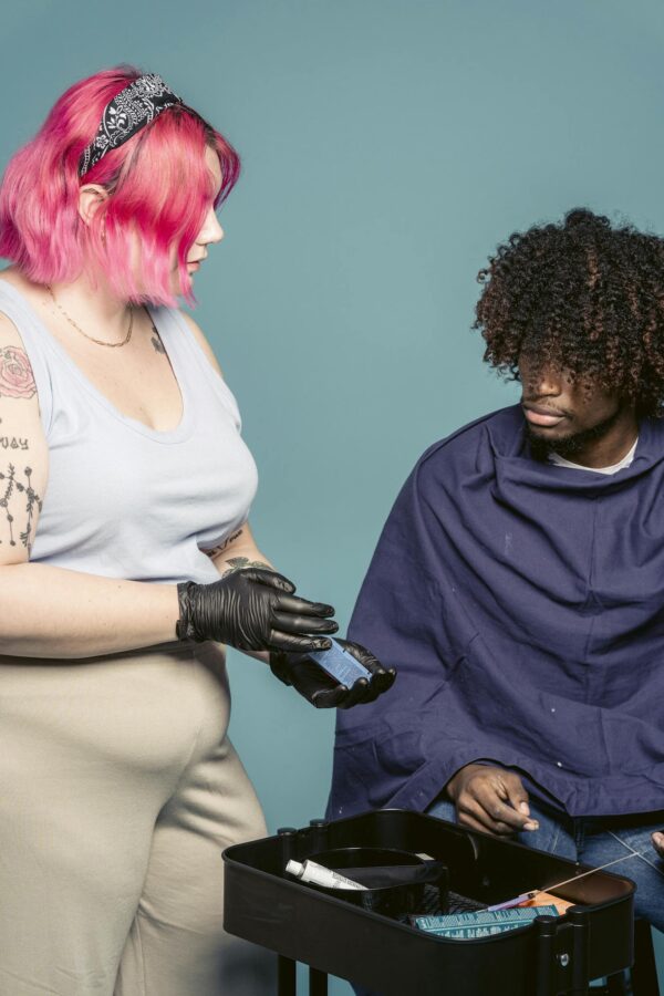 Young woman with pink hair demonstrating pigment to African American man against blue background in salon